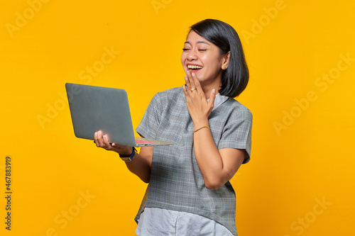 Happy young asian woman holding laptop laughing and covering mouth with hands isolated over yellow background