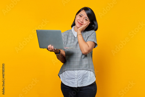 Portrait of cheerful attractive asian woman using laptop and touching cheeks over yellow background