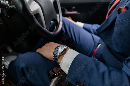 A man in a blue suit and red tie with a watch on his arm is driving a car.