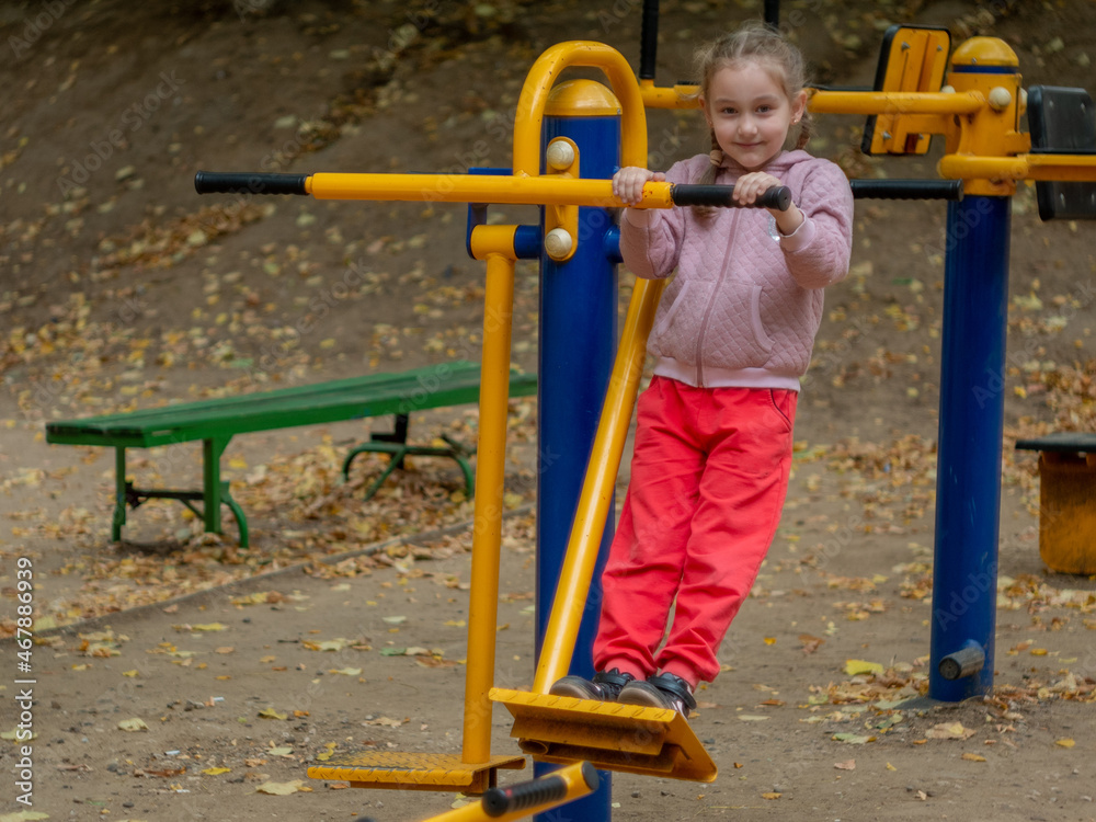 Sports ground in the city park. A girl of 7-8 years old is engaged in a sports simulator. Sports ground in the city park. A girl of 7-8 years old is engaged in a sports simulator.