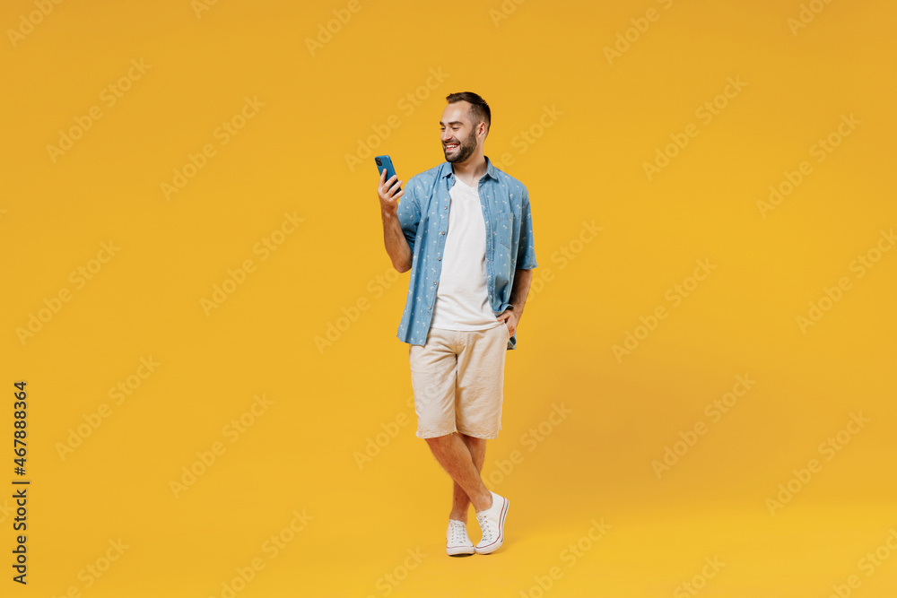 Full body young smiling happy cool caucasian man 20s wearing blue shirt white t-shirt hold in hand use mobile cell phone isolated on plain yellow background studio portrait. People lifestyle concept. - obrazy, fototapety, plakaty 