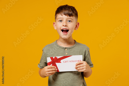Little small smiling happy boy 6-7 years old wearing green t-shirt hold gift certificate coupon voucher card for store isolated on plain yellow background. Mother's Day love family lifestyle concept.