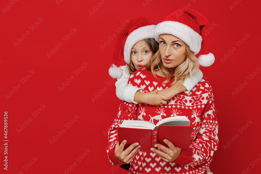 Young woman wear sweater hat have fun with child baby girl 6-7 years old. Mommy little kid daughter cuddle hug read book isolated on plain red color background studio. New Year love family concept Young woman wear sweater hat have fun with child baby girl 6-7 years old. Mommy little kid daughter cuddle hug read book isolated on plain red color background studio. New Year love family concept
