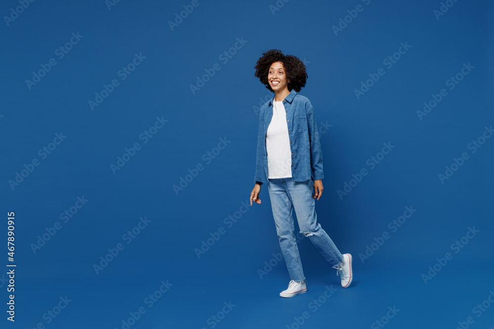 Full body side view fun young smiling happy black woman in casual clothes shirt white t-shirt walking going look aside isolated on plain dark blue background studio portrait. People lifestyle concept. Full body side view fun young smiling happy black woman in casual clothes shirt white t-shirt walking going look aside isolated on plain dark blue background studio portrait. People lifestyle concept.