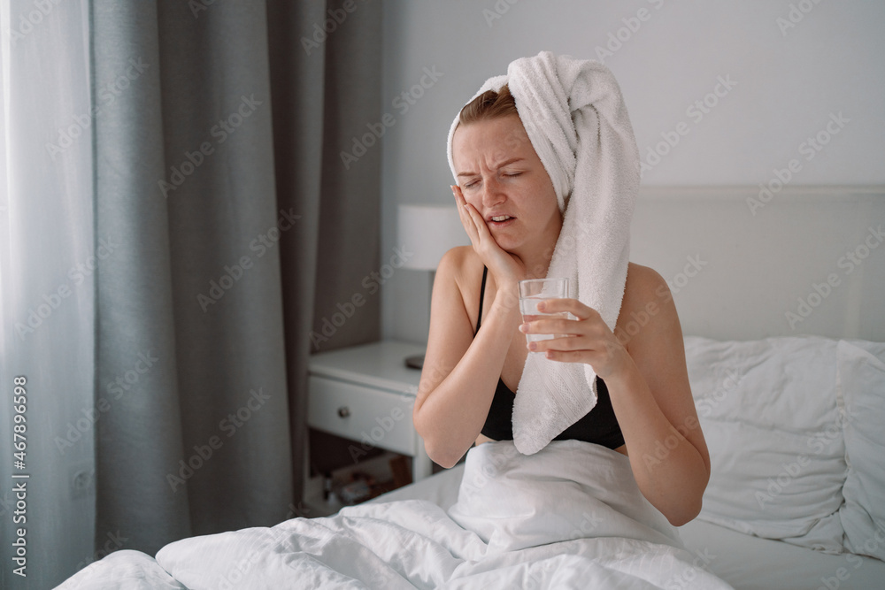 Sad unhappy europian girl with toothache holds a glass of clean water in a white bed of a bed at home in the morning