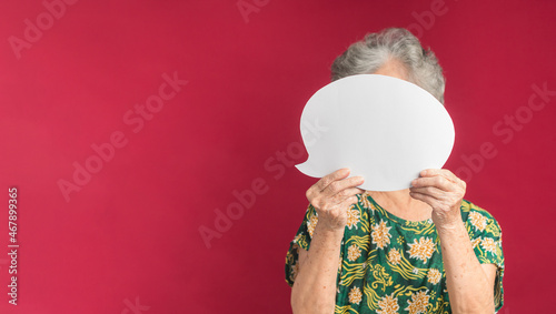 Concept of a speech bubble. A senior woman with short gray hair holding a blank white paper speech bubble closed her face while standing on a red background in the studio. Space for text
