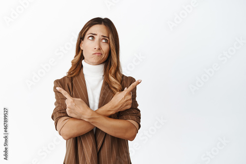 Portrait of indecisive woman pointing sideways with confused face, having doubts while making her choice, white background