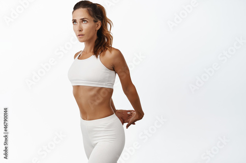 Middle aged woman with strong fit body, stretching arms behind, warm up and workout, standing over white background