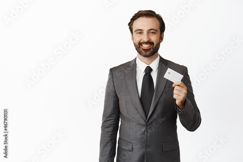 Handsome successful corporate man showing credit card and smiling happy, standing in suit against studio background