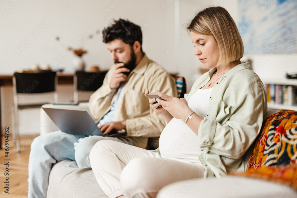Man and pregnant woman using cellphone and laptop while sitting on couch Man and pregnant woman using cellphone and laptop while sitting on couch