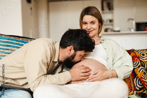 White man kissing his pregnant wife's belly while sitting on couch