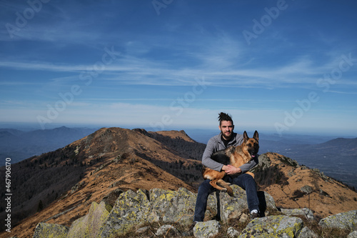 Two tourists travel together through national park - man and German Shepherd. Caucasian young man with dreadlocks sits on top of mountain and hugs his dog.