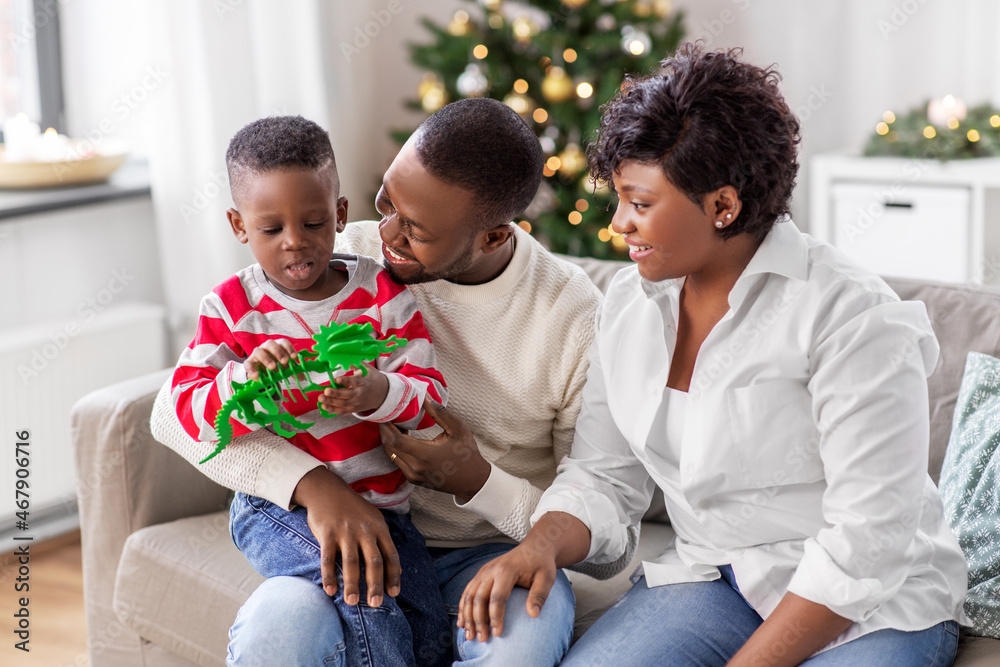 family, winter holidays and people concept - happy african american mother, father and baby son with dinosaur toy at home on christmas