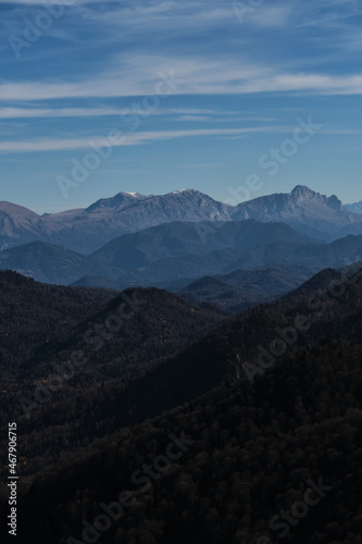 Beautiful panoramic landscape view of mountains on warm sunny autumn day. Blue sky and white clouds. In distance, steep cliffs and silhouettes of large snow capped mountain peaks.