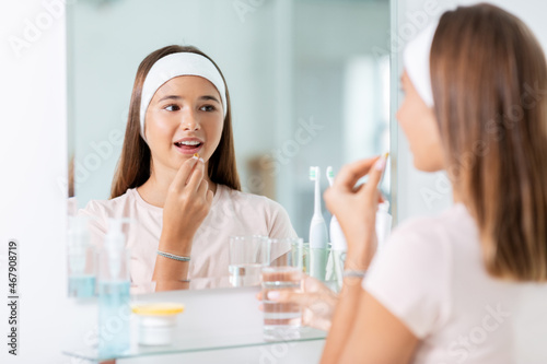 beauty, hygiene and people concept - happy teenage girl with cod liver oil capsule and glass of water looking in mirror at bathroom