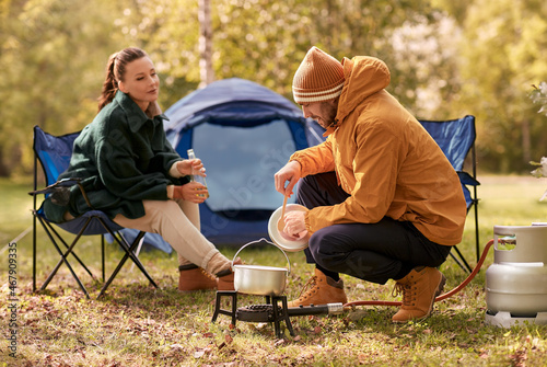 camping, tourism and travel concept - happy couple drinking beer and cooking food in pot on tourist gas burner at tent camp photo