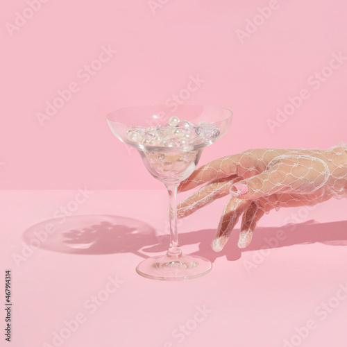 Creative layout with hand in white lace glove  raeching martini cocktail glass with diamonds and pearls on pastel pink background. 80s or 90s retro aesthetic fashion concept. Romantic drink idea.