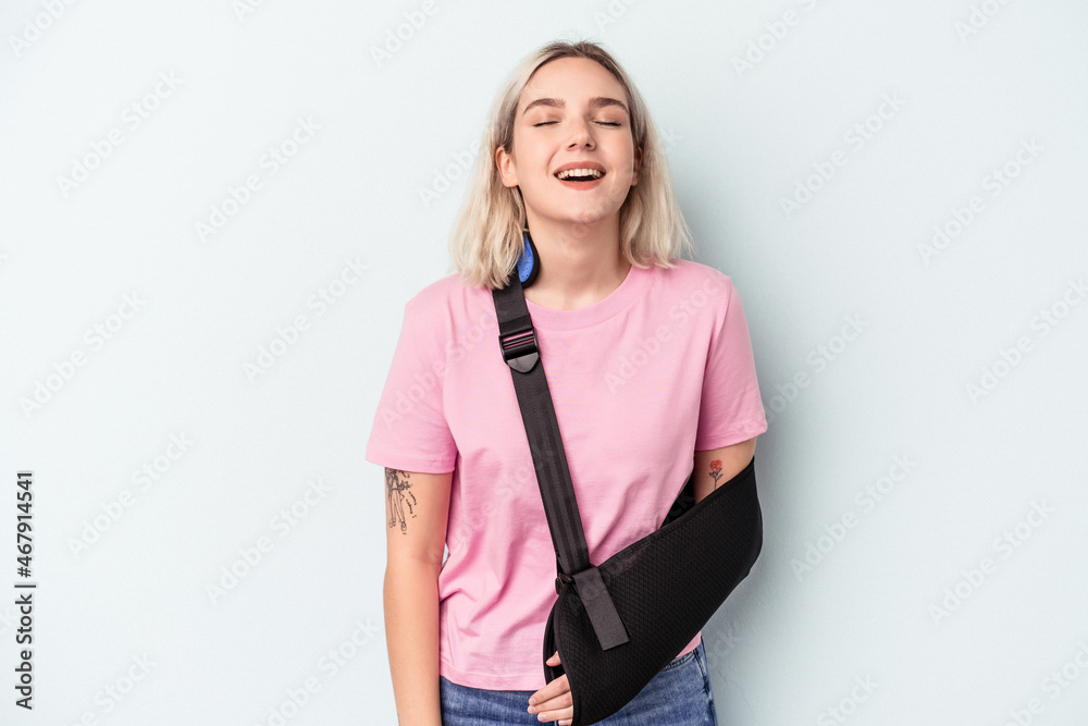 Young caucasian woman with broken hand isolated on blue background laughing and having fun.