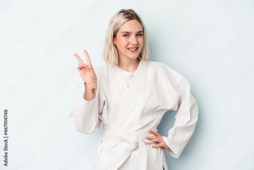 Fototapeta premium Young caucasian woman doing karate isolated on blue background joyful and carefree showing a peace symbol with fingers.