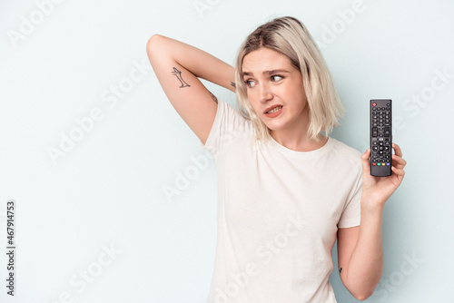 Young caucasian woman holding a tv controller isolated on blue background touching back of head, thinking and making a choice.