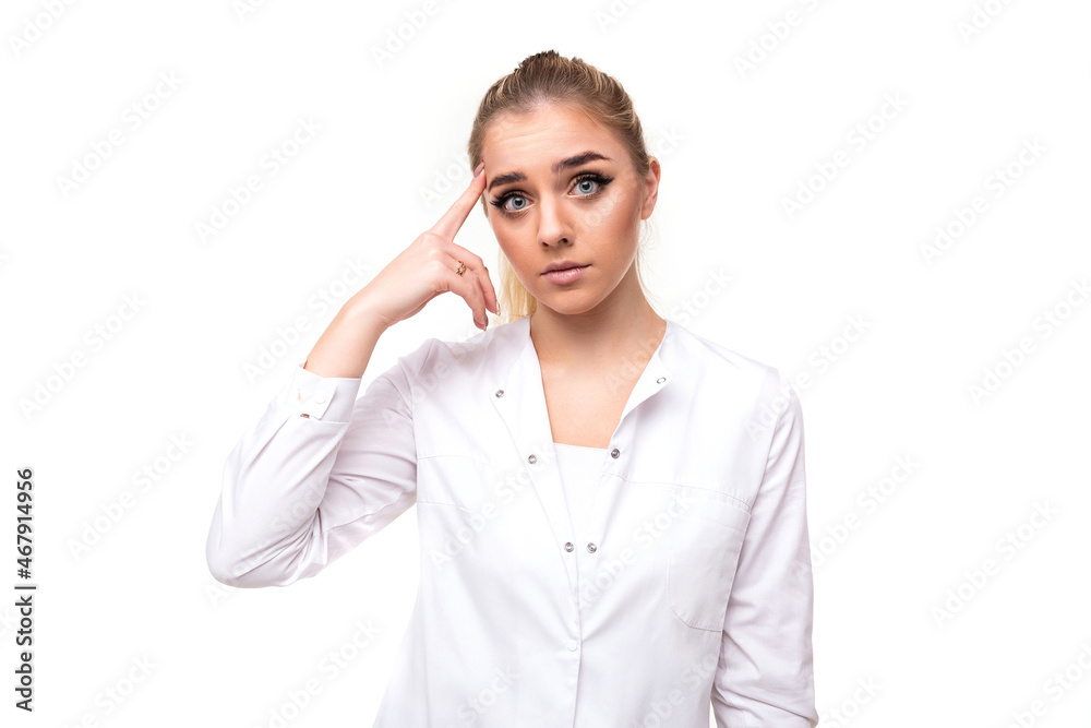 Photo of an attractive sad young blonde woman with a ponytail and makeup in a white medical coat of a nurse touches herself with finger isolated on a white background with free and copy space for text Photo of an attractive sad young blonde woman with a ponytail and makeup in a white medical coat of a nurse touches herself with finger isolated on a white background with free and copy space for text