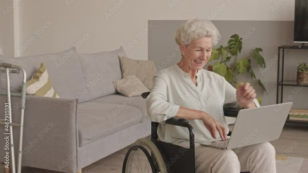 Medium stab shot of smiling Caucasian senior woman sitting in wheelchair at cozy living room having video conversation on laptop Medium stab shot of smiling Caucasian senior woman sitting in wheelchair at cozy living room having video conversation on laptop