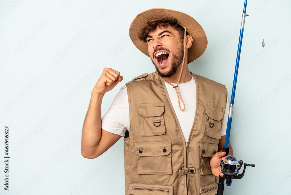 Young mixed race fisherman holding a rod isolated on blue background raising fist after a victory, winner concept. Young mixed race fisherman holding a rod isolated on blue background raising fist after a victory, winner concept.
