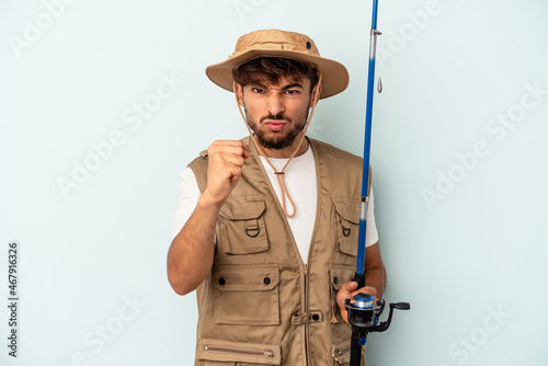 Young mixed race fisherman holding a rod isolated on blue background showing fist to camera, aggressive facial expression.