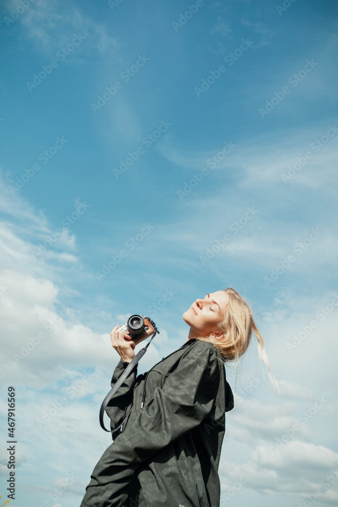 Young blond woman in khaki shirt with a hood holding camera in front of blue sky outdoors Young blond woman in khaki shirt with a hood holding camera in front of blue sky outdoors
