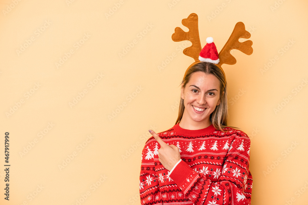 Young caucasian woman wearing a christmas reindeer hat isolated on yellow background smiling and pointing aside, showing something at blank space. Young caucasian woman wearing a christmas reindeer hat isolated on yellow background smiling and pointing aside, showing something at blank space.