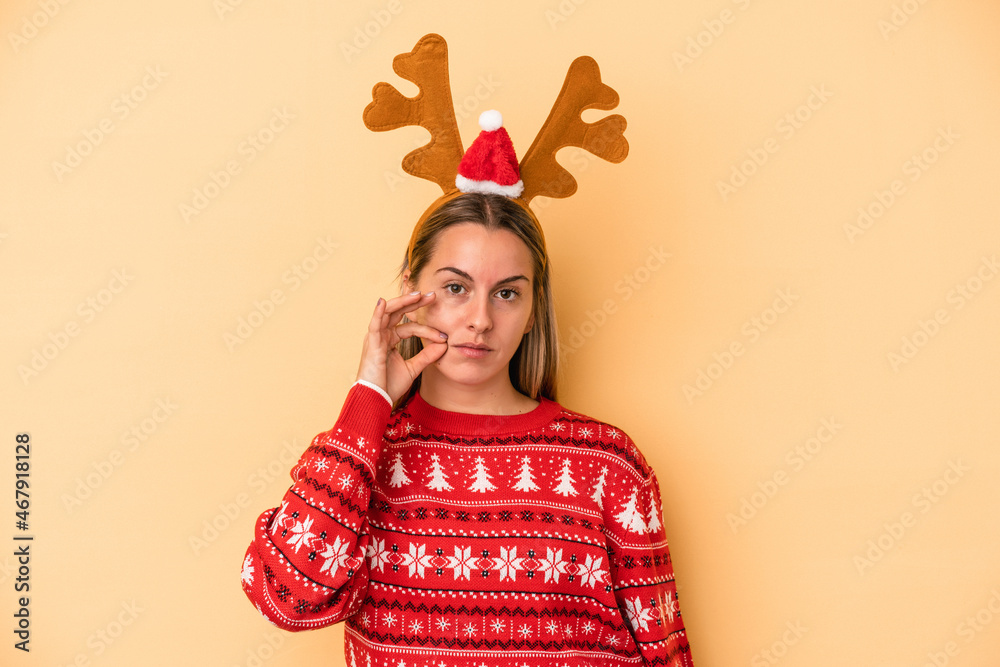 Young caucasian woman wearing a christmas reindeer hat isolated on yellow background with fingers on lips keeping a secret. Young caucasian woman wearing a christmas reindeer hat isolated on yellow background with fingers on lips keeping a secret.