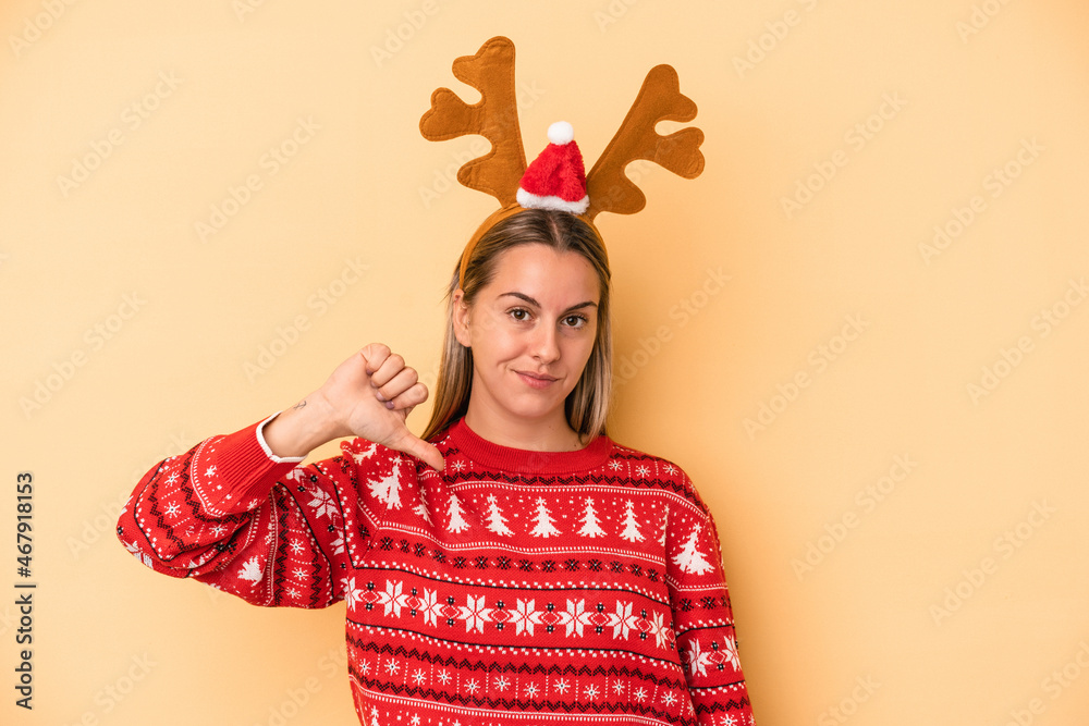 Young caucasian woman wearing a christmas reindeer hat isolated on yellow background showing a dislike gesture, thumbs down. Disagreement concept. Young caucasian woman wearing a christmas reindeer hat isolated on yellow background showing a dislike gesture, thumbs down. Disagreement concept.