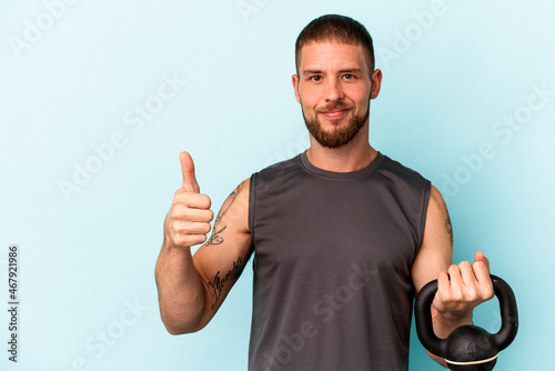 Young caucasian man holding ketlebell isolated on blue background smiling and raising thumb up photo