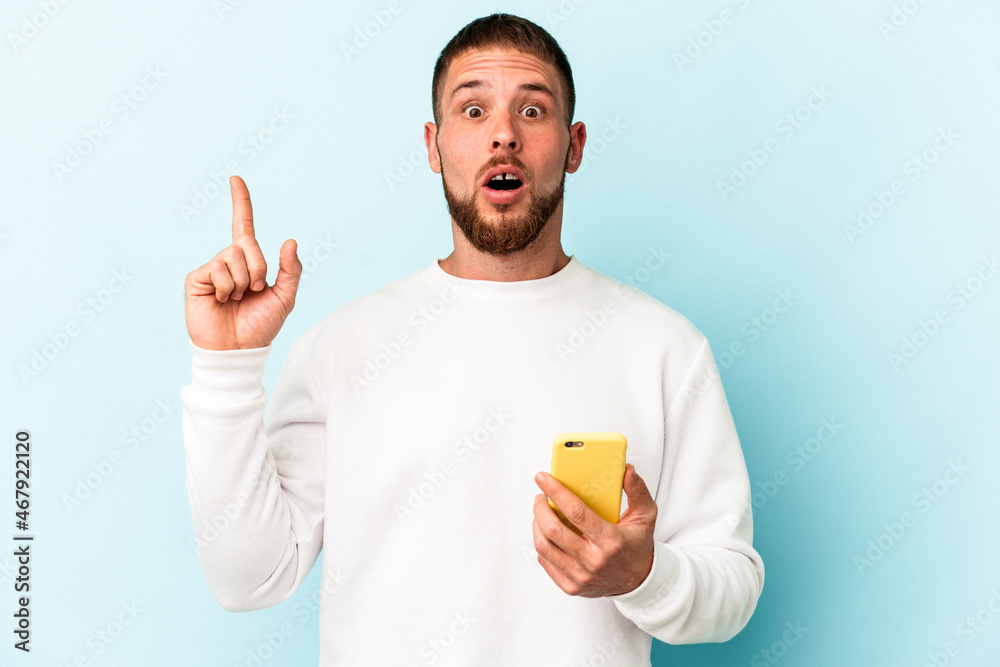 Young caucasian man holding mobile phone isolated on blue background having an idea, inspiration concept. Young caucasian man holding mobile phone isolated on blue background having an idea, inspiration concept.