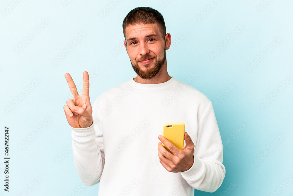 Young caucasian man holding mobile phone isolated on blue background showing number two with fingers. Young caucasian man holding mobile phone isolated on blue background showing number two with fingers.