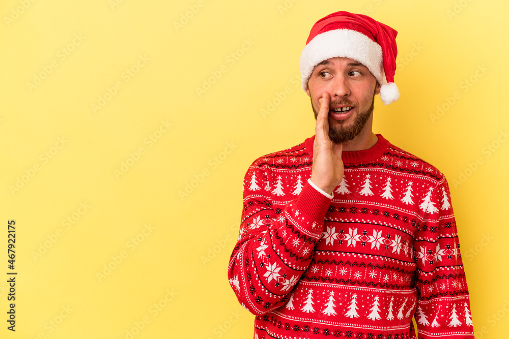 Young caucasian man celebrating Christmas isolated on yellow background is saying a secret hot braking news and looking aside