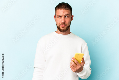 Young caucasian man holding mobile phone isolated on blue background confused, feels doubtful and unsure.