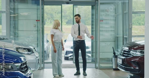 Female customer walking with salesman at modern salon and observing various luxury auto. Competent dealer helping young woman to make expensive purchase.