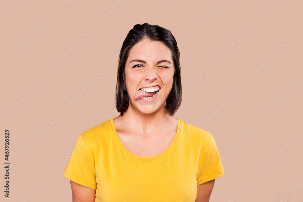 Close up beautiful cheerful woman making a funny facial expression, with one eye closed and sticking her tongue out to the side. Young woman wearing yellow shirt over beige background.