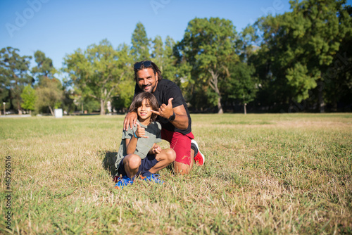 Happy father with disability with son. Man with mechanical leg in sunglasses and little boy sitting on grass in park, showing cool sign with fingers. Disability, family, love concept