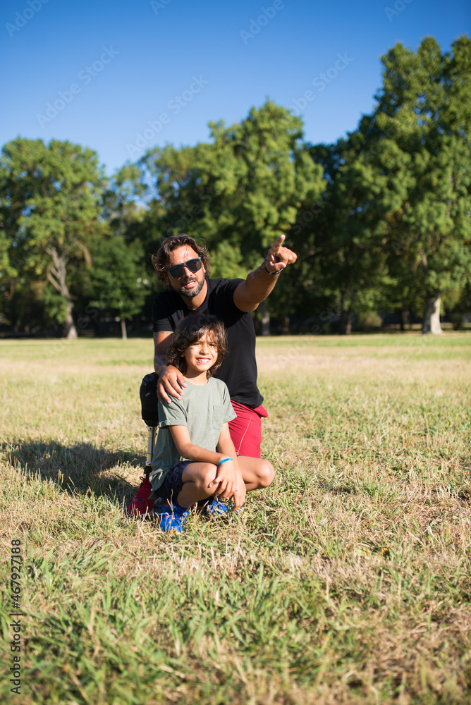 Obraz premium Loving father with disability talking with son. Man with mechanical leg in sunglasses and little boy sitting on grass in park, showing bird to son. Disability, family, love concept