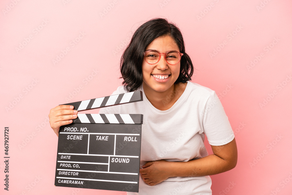 Young latin woman holding clapperboard isolated on pink background laughing and having fun.