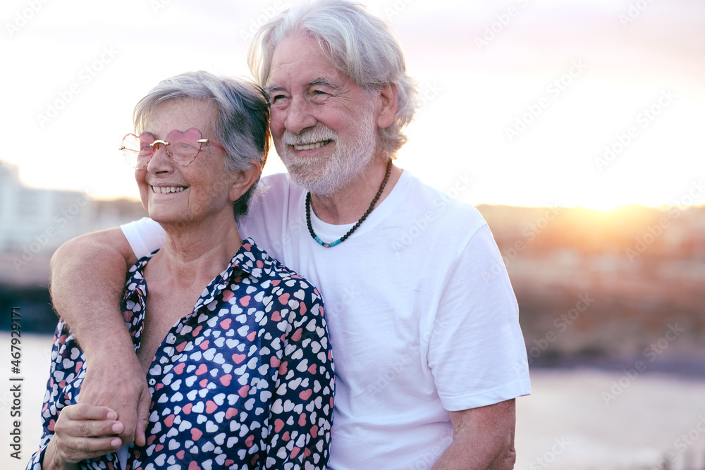 Beautiful caucasian senior couple smiling in outdoor at sea looking away. Elderly couple white haired enjoying retirement and sunset light
