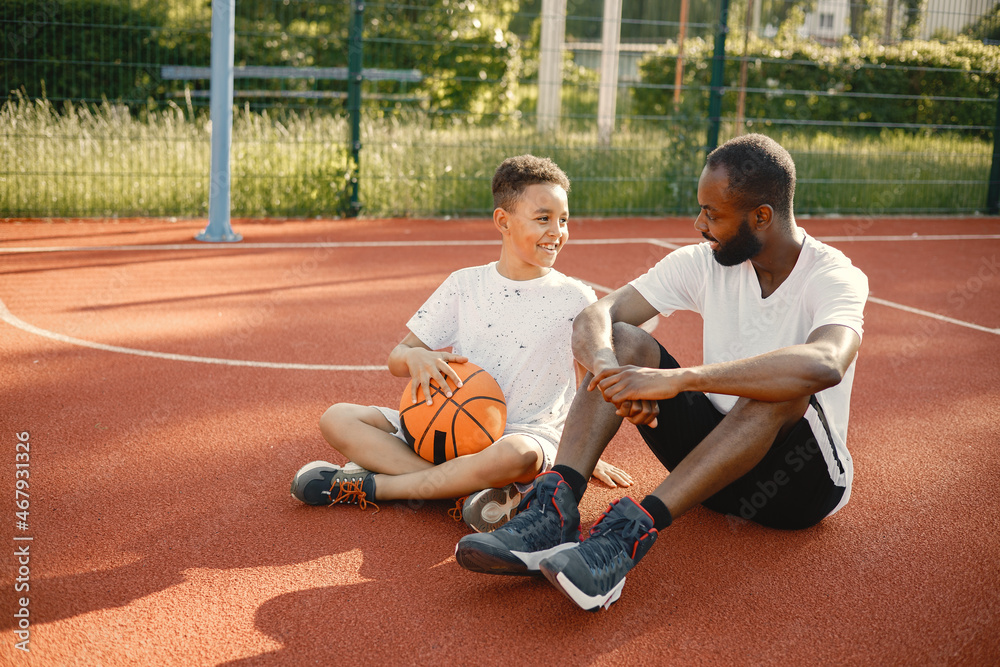 Black father with his multiracial son sitting on a basketball court together Black father with his multiracial son sitting on a basketball court together