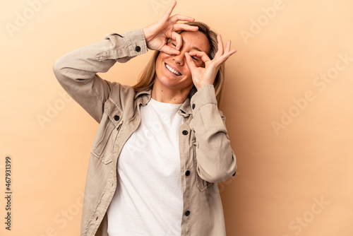 Young caucasian woman isolated on beige background showing okay sign over eyes