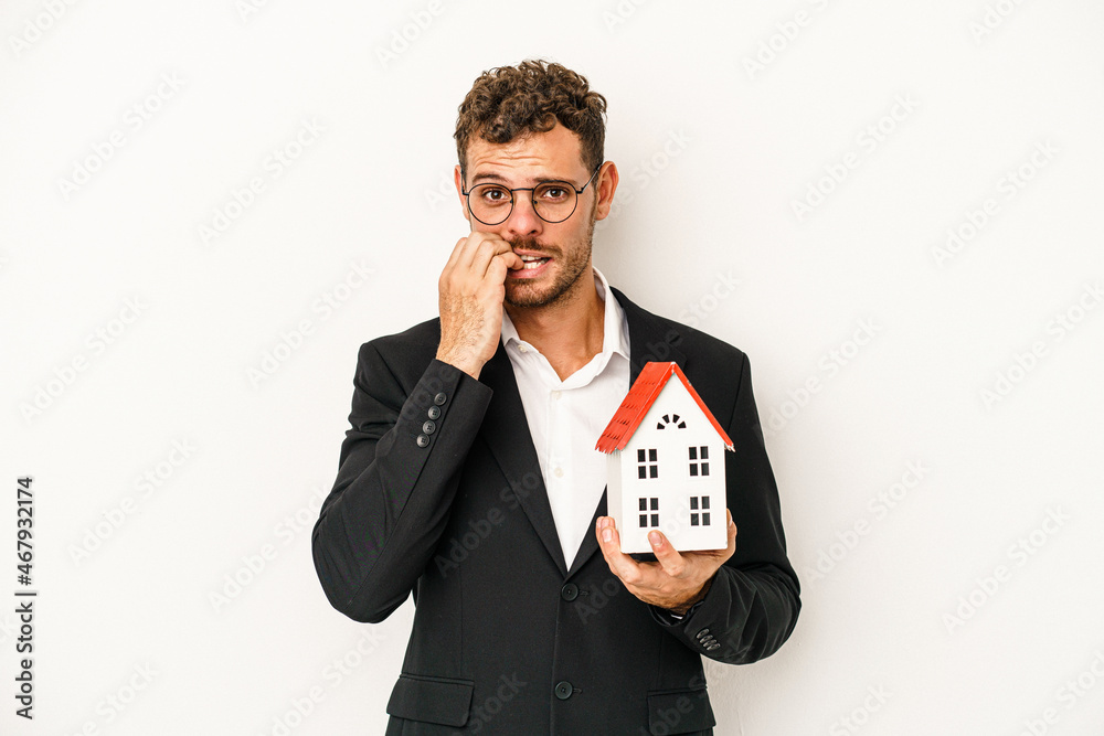Young caucasian real estate holding a toy home isolated on white background biting fingernails, nervous and very anxious.