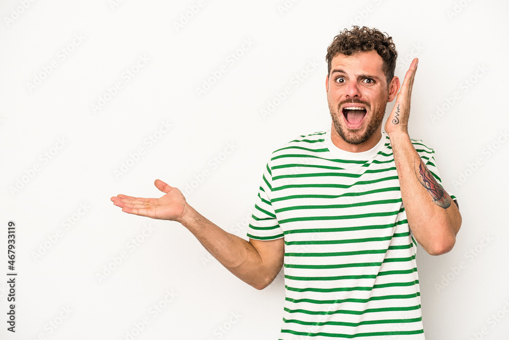 Young caucasian man isolated on white background holds copy space on a palm, keep hand over cheek. Amazed and delighted.