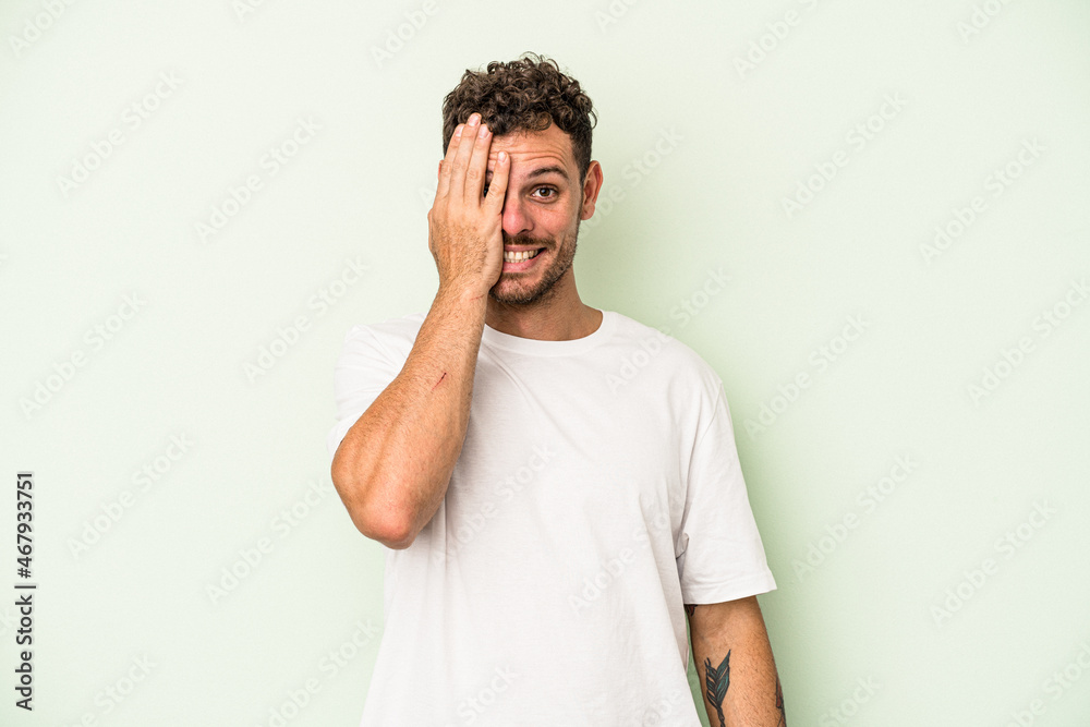 Young caucasian man isolated on green background having fun covering half of face with palm. Young caucasian man isolated on green background having fun covering half of face with palm.