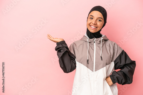 Young Arab woman with sport burqa isolated on pink background showing a copy space on a palm and holding another hand on waist.