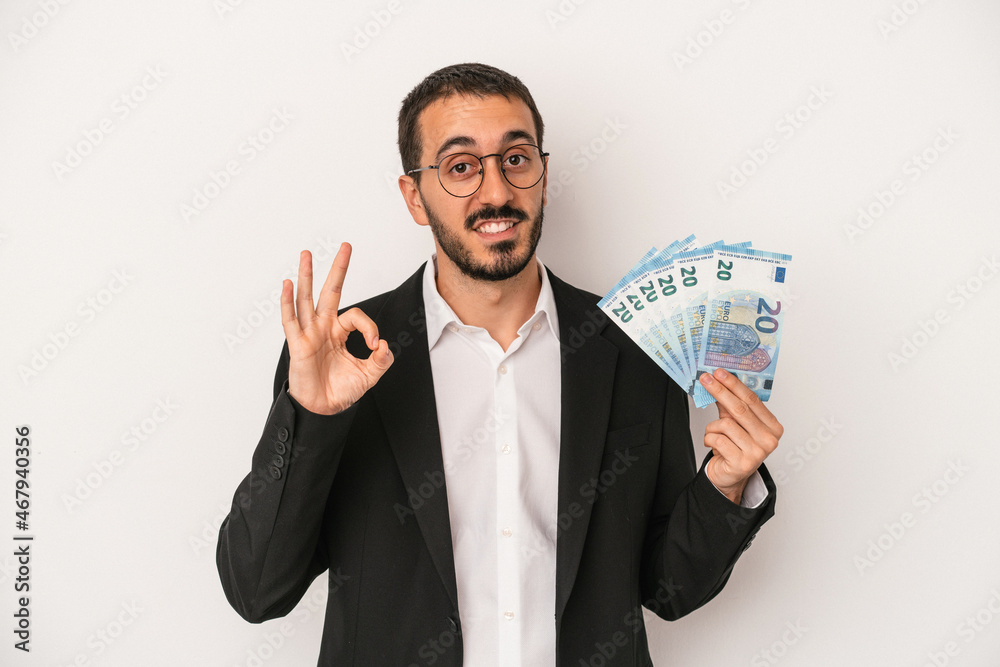 Young caucasian business man holding banknotes isolated on white background cheerful and confident showing ok gesture. Young caucasian business man holding banknotes isolated on white background cheerful and confident showing ok gesture.